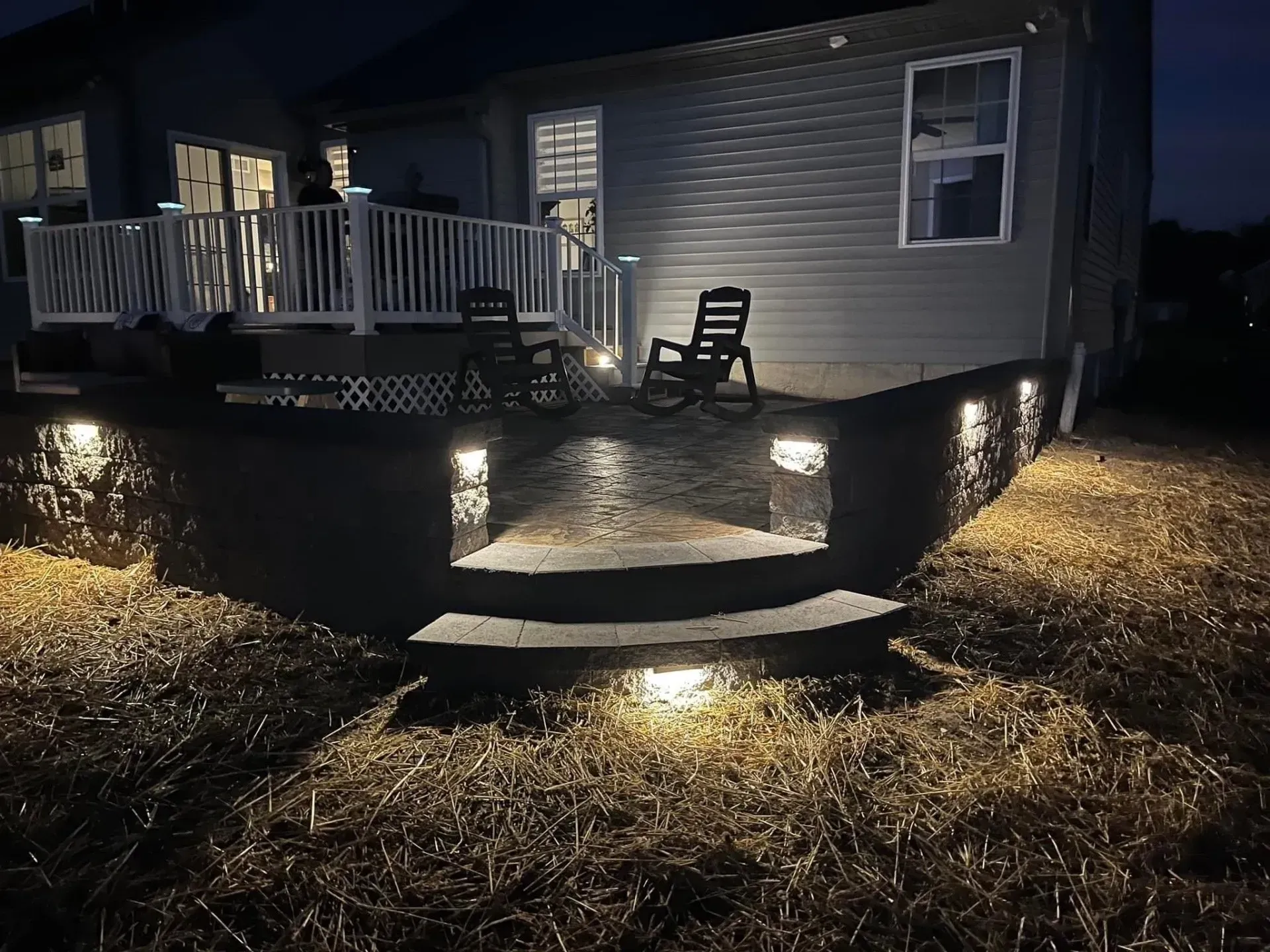 Backyard deck at night with built-in lighting on retaining walls and steps. Two rocking chairs sit on the deck.
