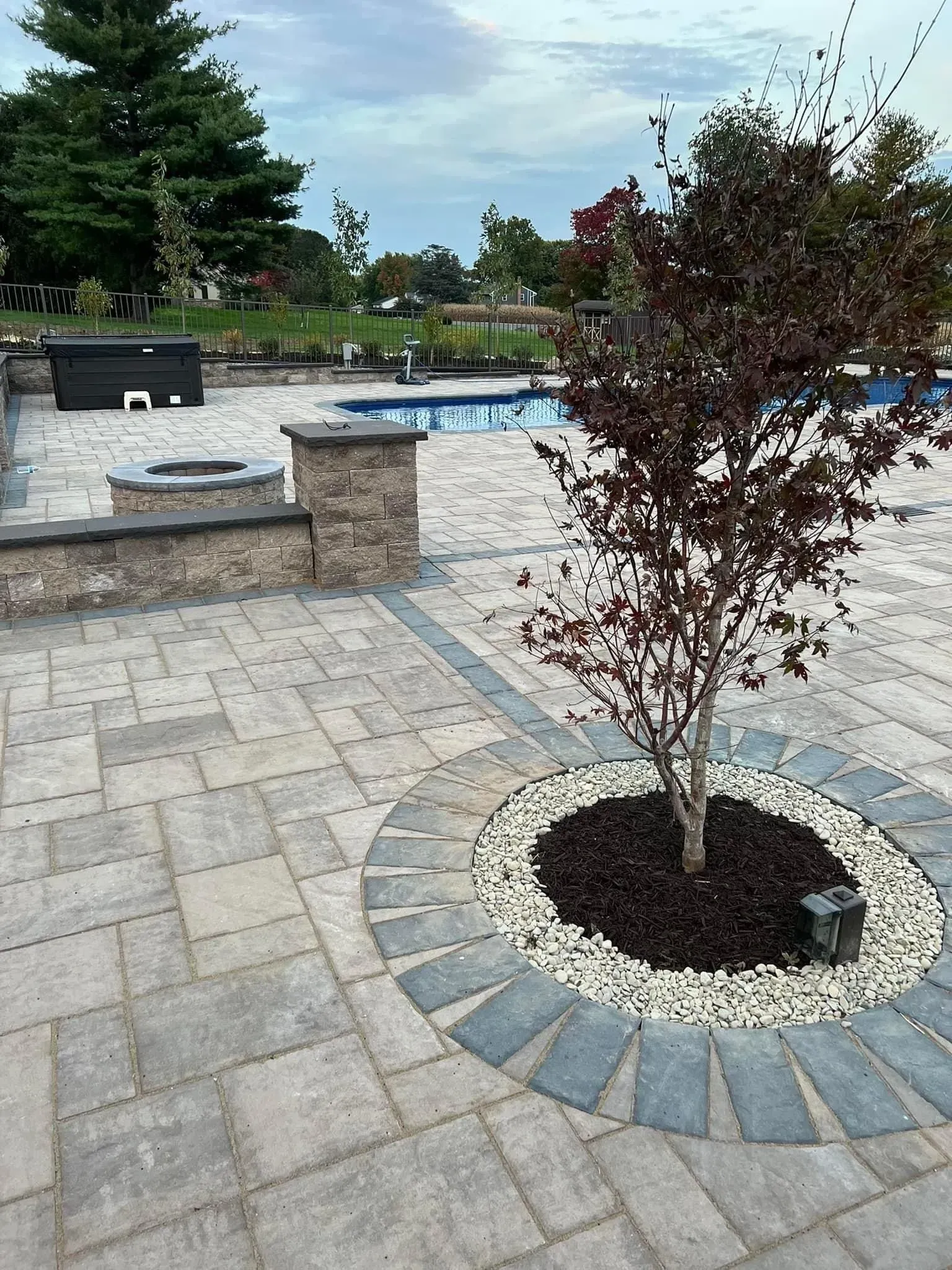Patio with a tree surrounded by stones, fire pit, pool in background.