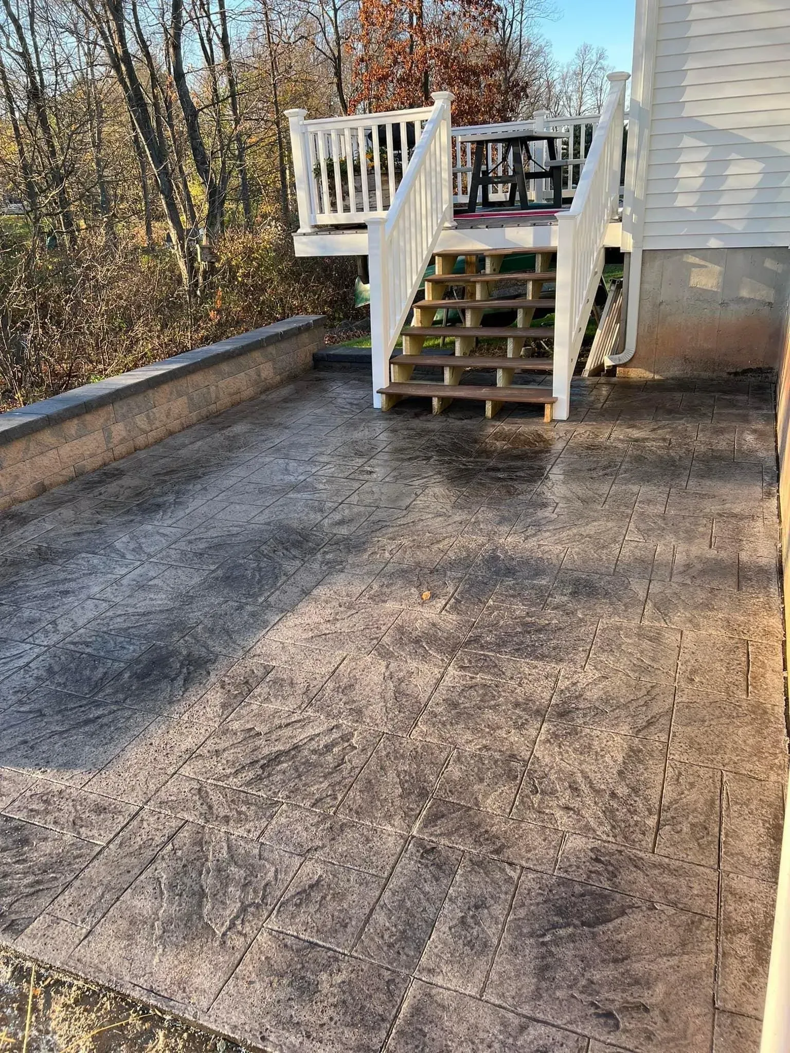 Stamped concrete patio with steps leading to a white deck, surrounded by trees.