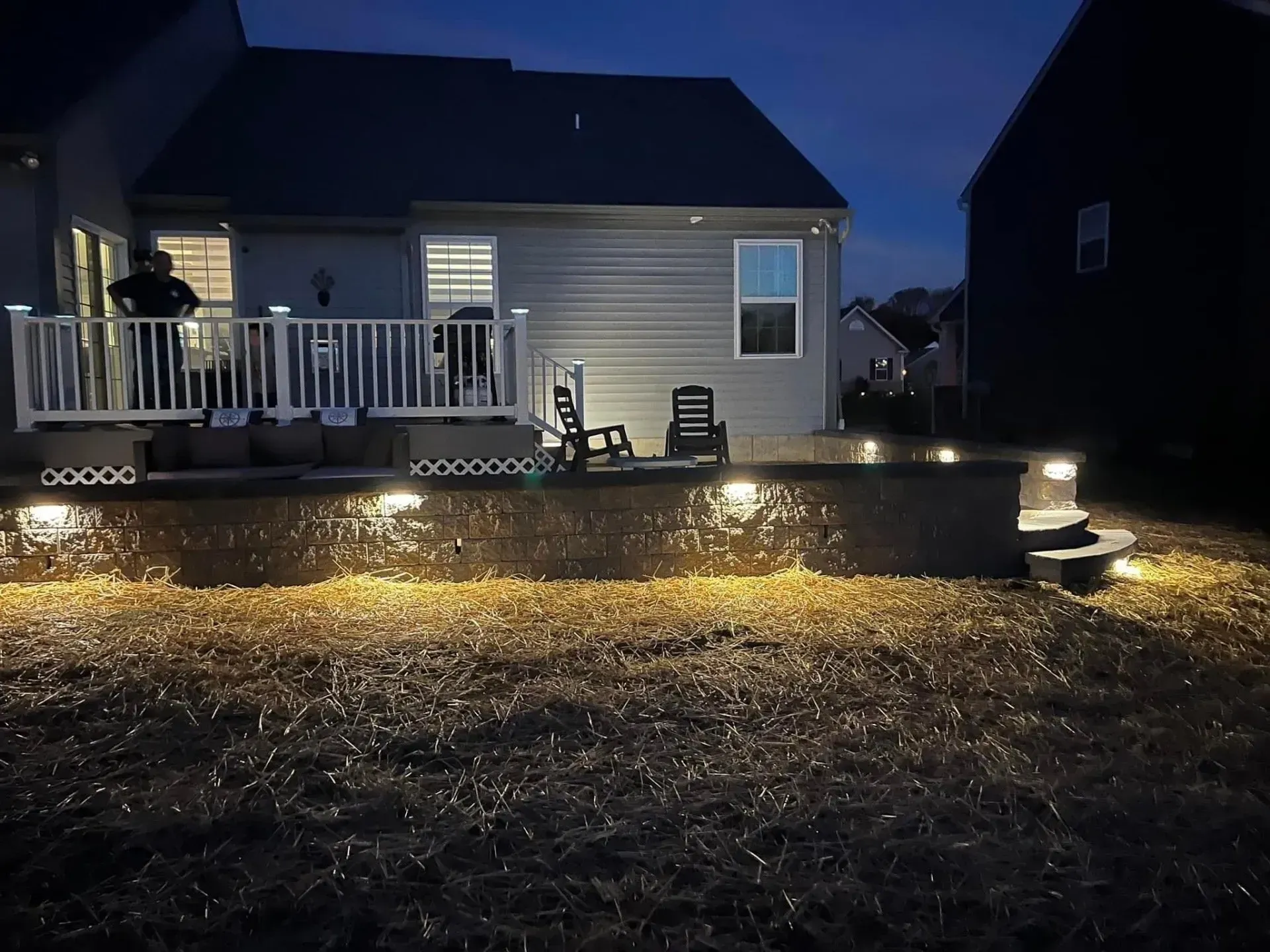 Backyard at dusk, house with deck, retaining wall with lights, and illuminated lawn.
