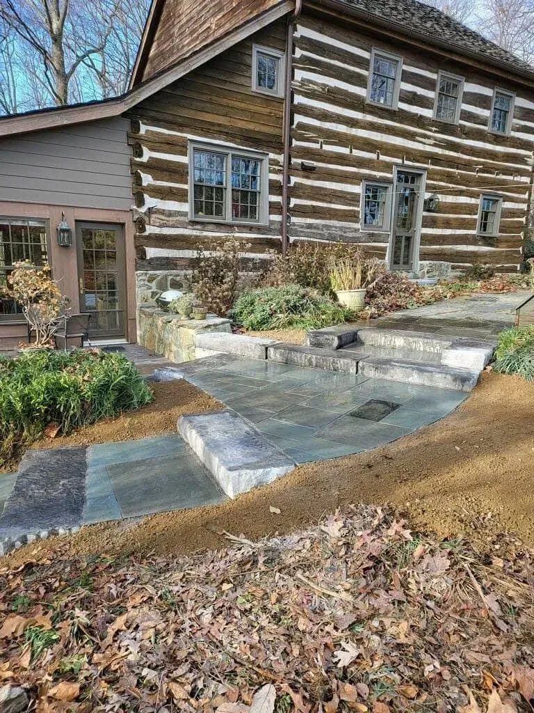 Log cabin house with stone patio and landscaping. Brown logs, gray stone, dry leaves on ground.