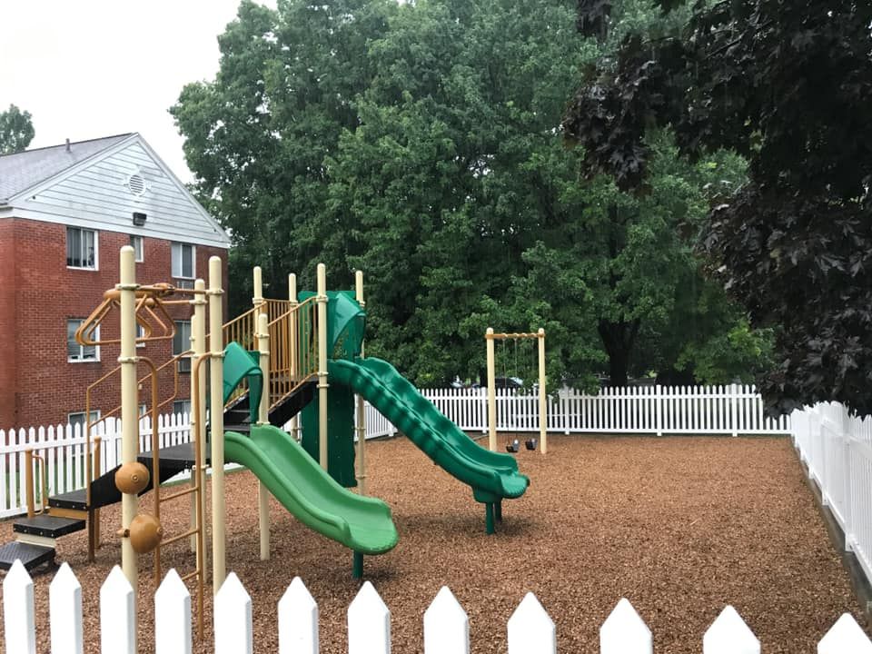 Playground with green slides, brown mulch, and white picket fence near a brick building and trees.