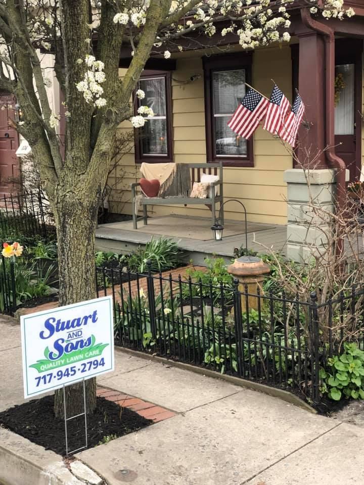 A yellow house with a porch, bench, American flags, and a yard with a black fence. A sign for Stuart & Sons.