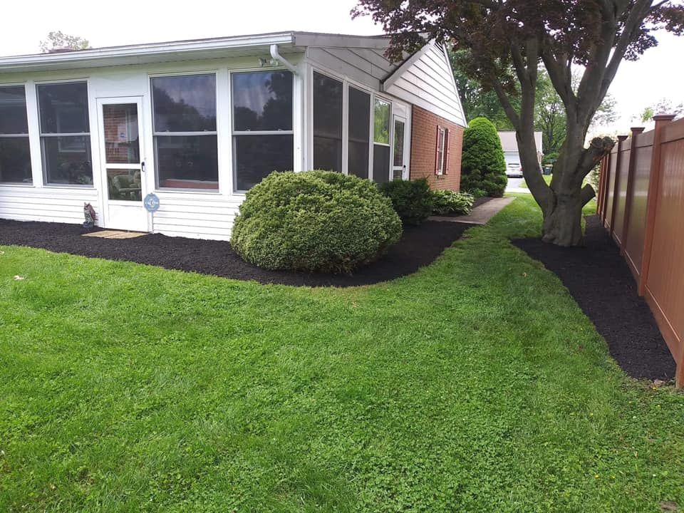 A manicured lawn with black mulch bordering a house and tree, next to a brown fence.