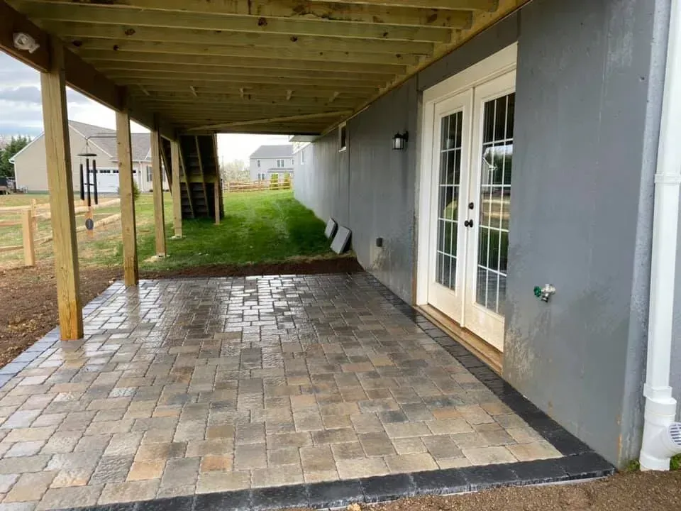 Patio under a deck with brick pavers, French doors, and a gray exterior wall.