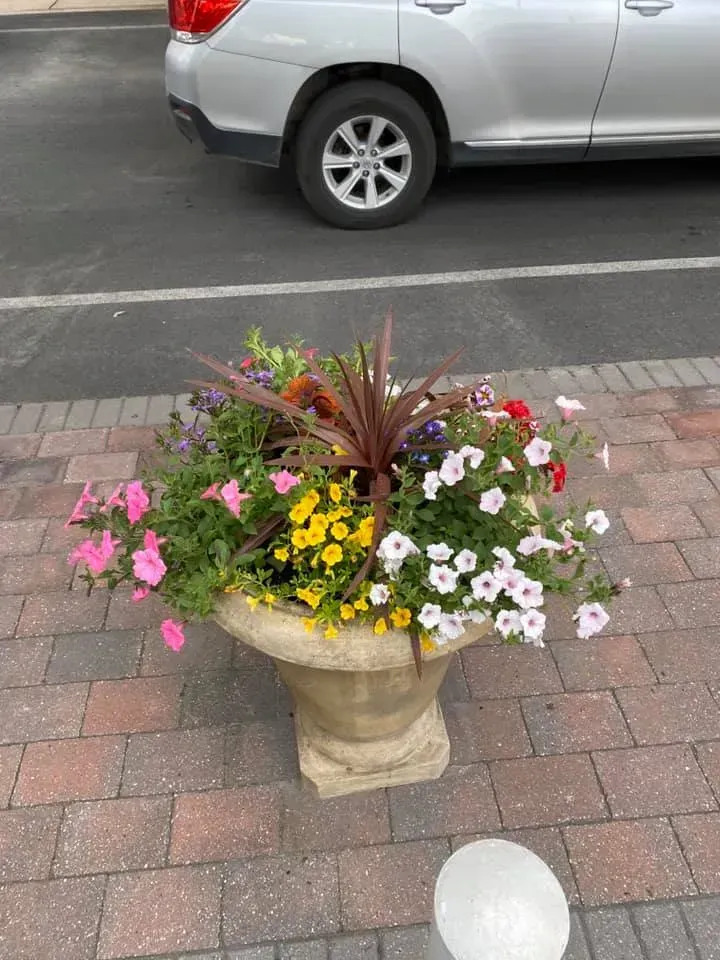 Stone planter overflowing with colorful flowers next to a silver car parked on a brick-paved area.