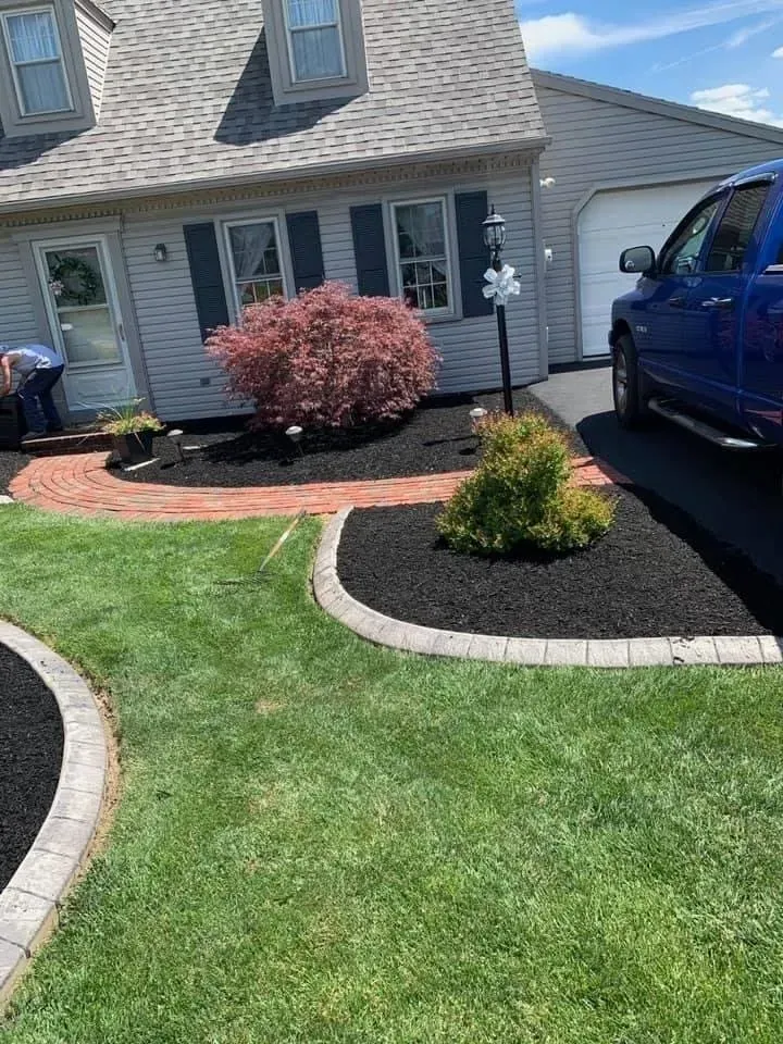 A house with a newly mulched garden bed, red brick edging, and green lawn. A blue truck is parked nearby.