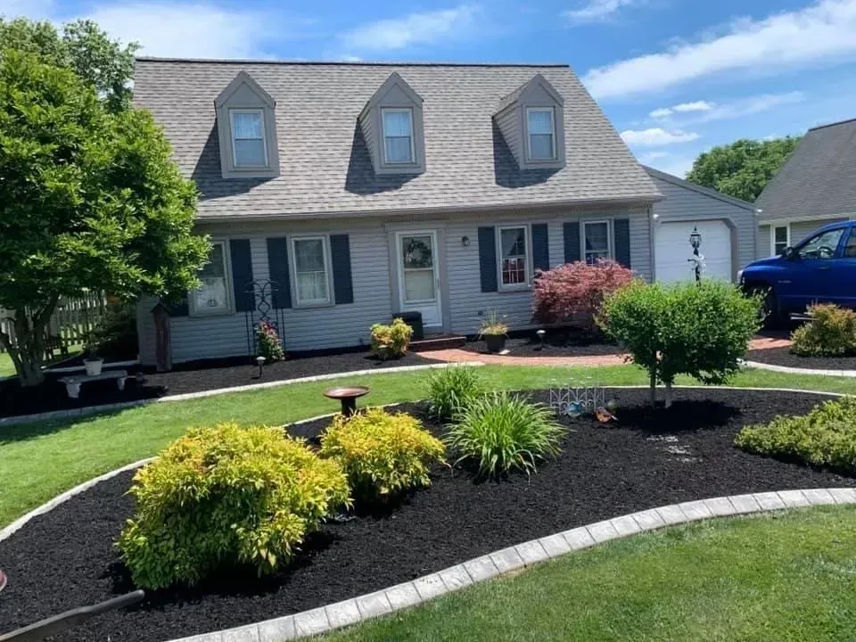 House with gray siding, dormers, and blue shutters, landscaped with dark mulch and green plants.