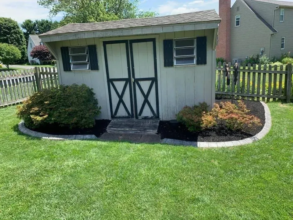 Wooden shed with green trim, black doors, small windows, and landscaping in a yard.