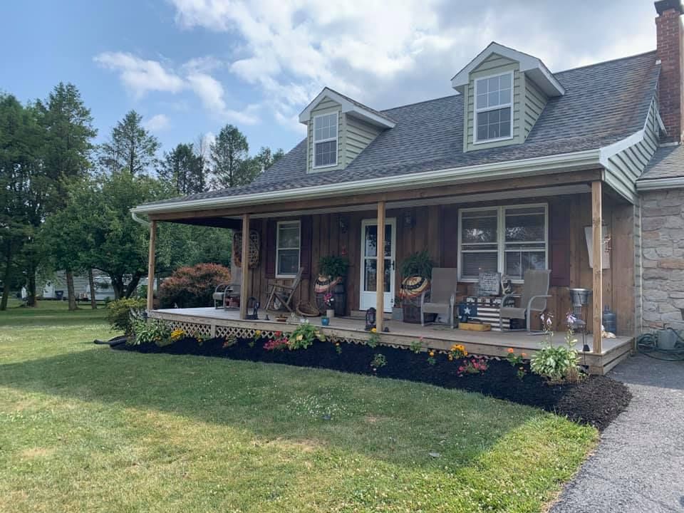 House with porch, landscaping, and dormer windows on a sunny day. Green lawn, trees, and dark mulch are visible.