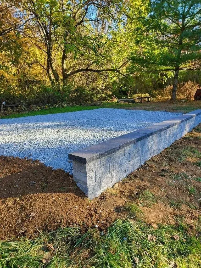 A low retaining wall of gray blocks borders a gravel area in a wooded setting.