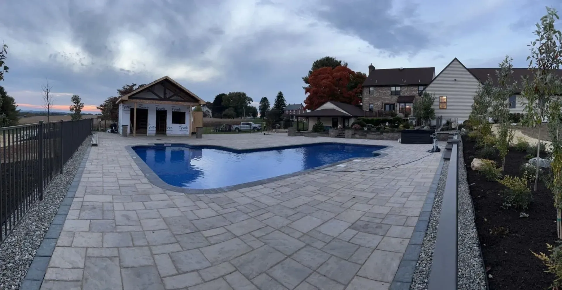 Pool with stone patio, fencing, and a house under an overcast sky.