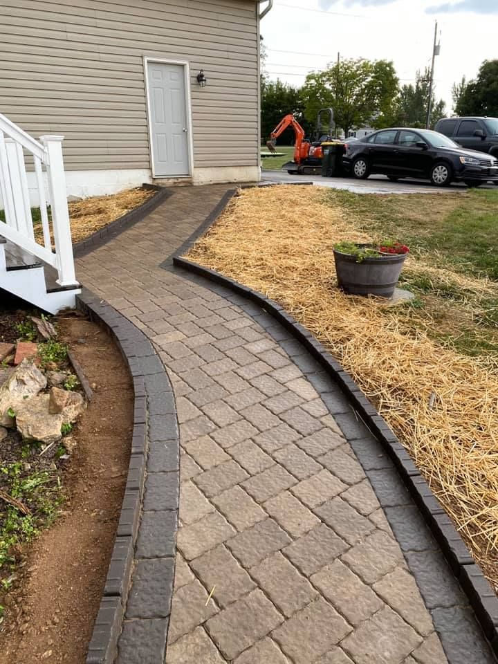 Brick pathway curves to a white door, bordered by dark pavers. Beige siding, lawn, and cars in background.