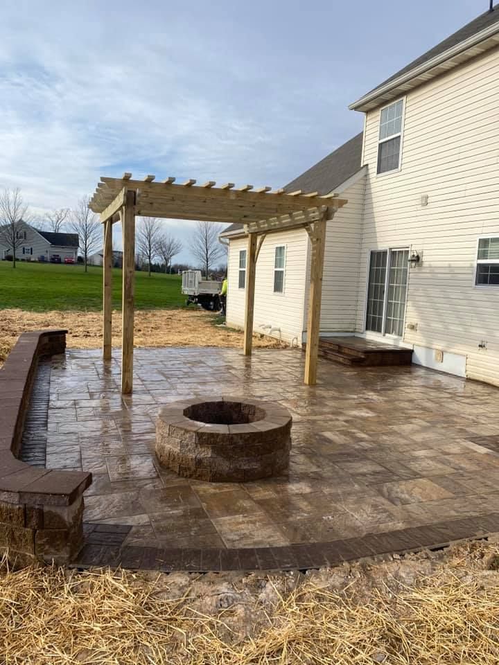 A backyard patio with a pergola, fire pit, and brickwork, next to a two-story house under a cloudy sky.