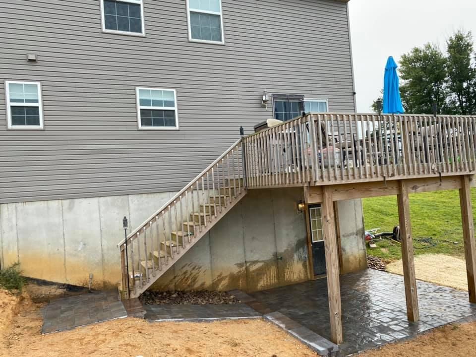 Wooden deck with stairs leading down to a concrete patio, next to a two-story house with gray siding.