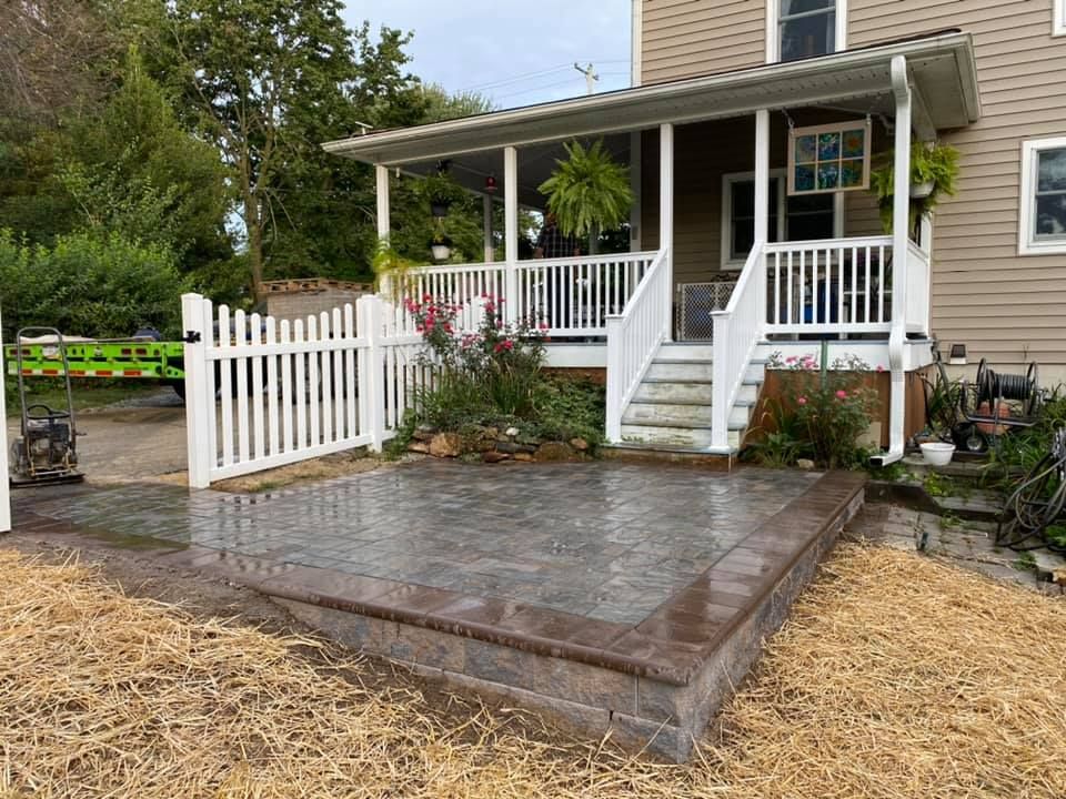 A new brick patio in front of a house, with a white picket fence and porch.