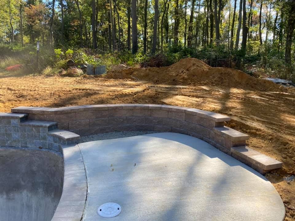 Concrete patio with curved retaining wall and steps, surrounded by dirt and trees.