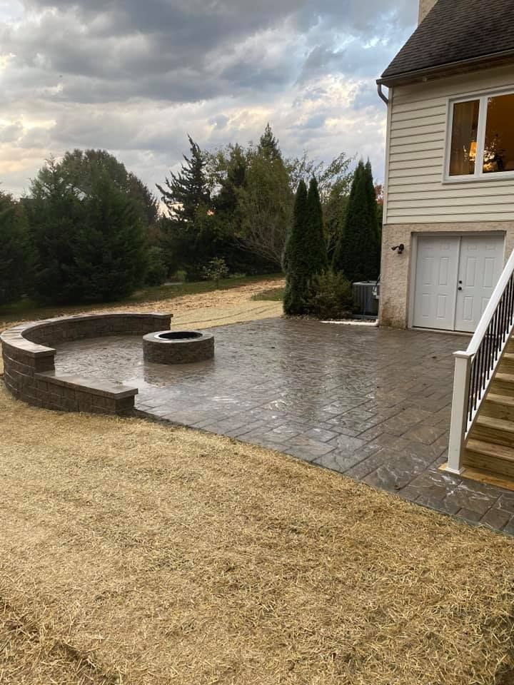 A paved patio with a fire pit, next to a beige house with a garage and stairs, under a cloudy sky.