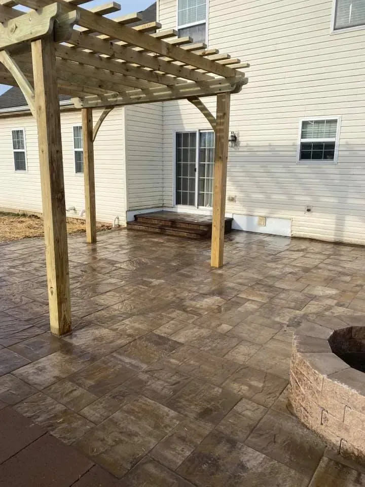 Wooden pergola over a brick patio next to a house with a sliding door and fire pit.