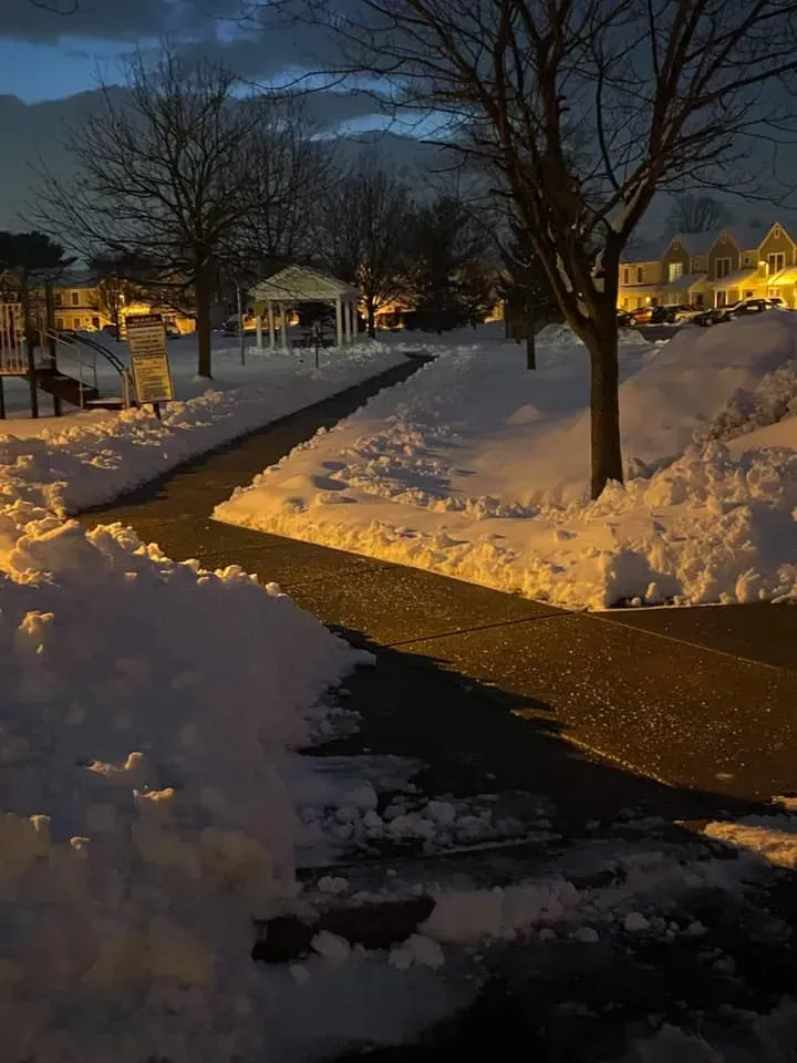 Snowy path at dusk, lit by streetlights. Park with bare trees, gazebo, and snow banks.
