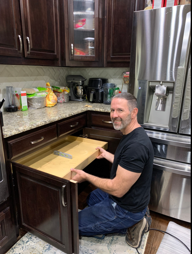 Man installing a drawer in a dark wood kitchen cabinet. He's kneeling, smiling, near a stainless steel refrigerator.