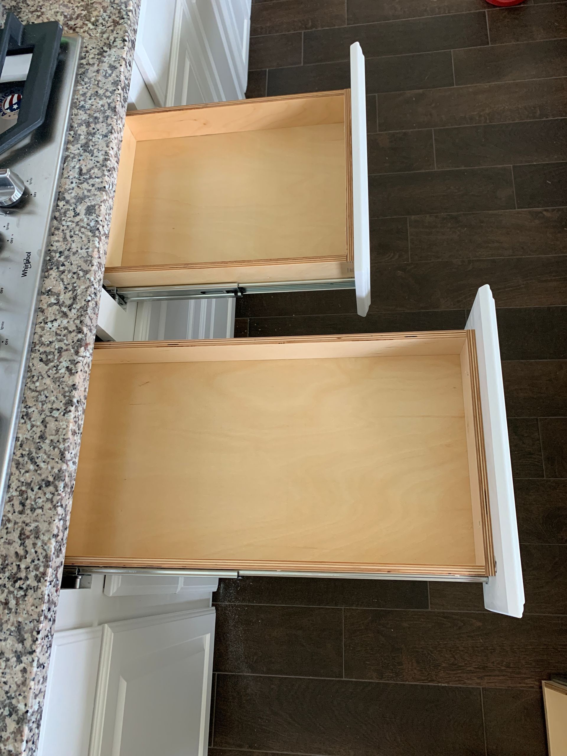 Two open kitchen drawers; white cabinets, wooden interiors. Drawers are empty, on a dark tiled floor.