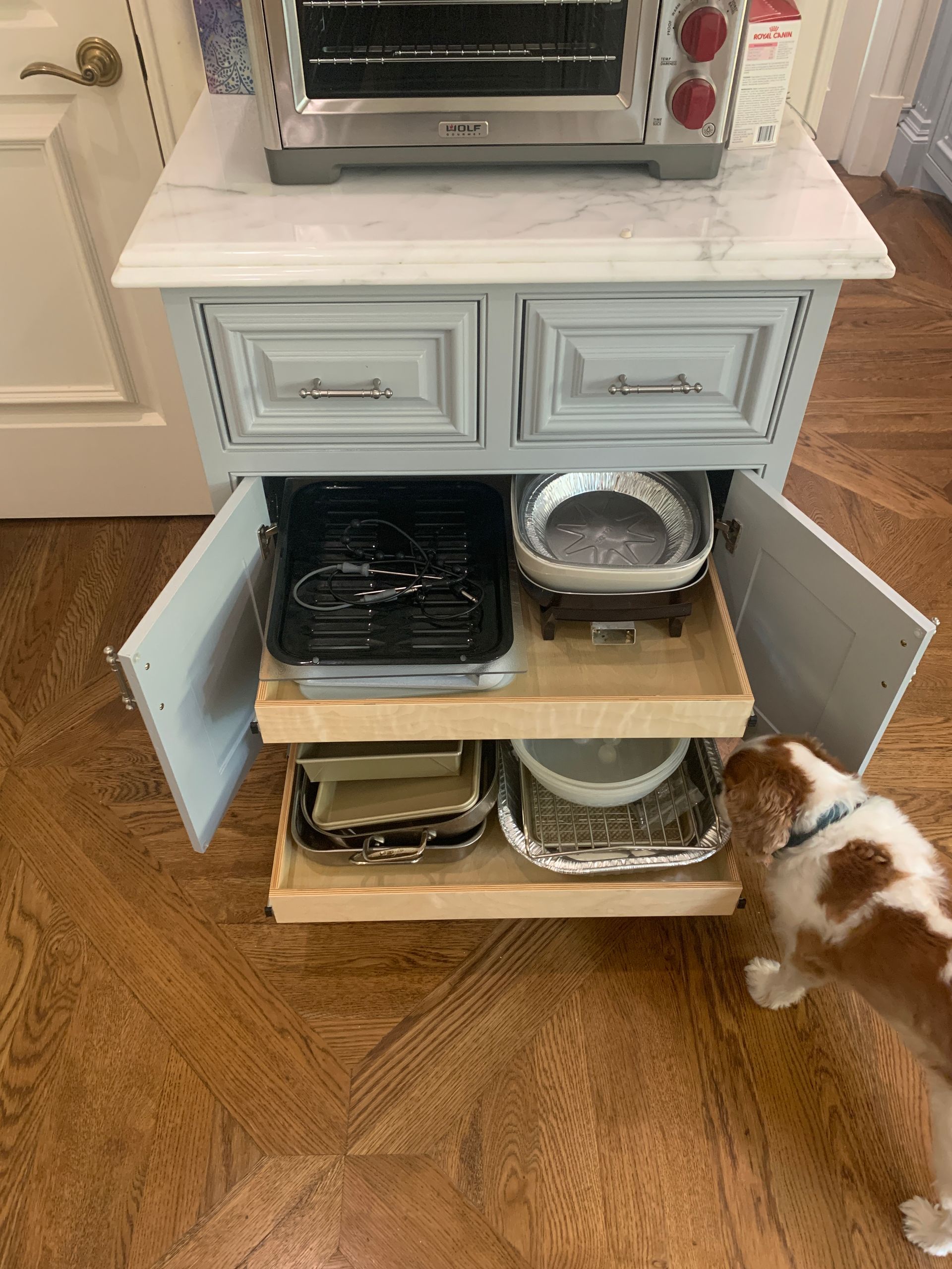 A small, blue and white cabinet with pull-out shelves holding various baking dishes, and a small dog.