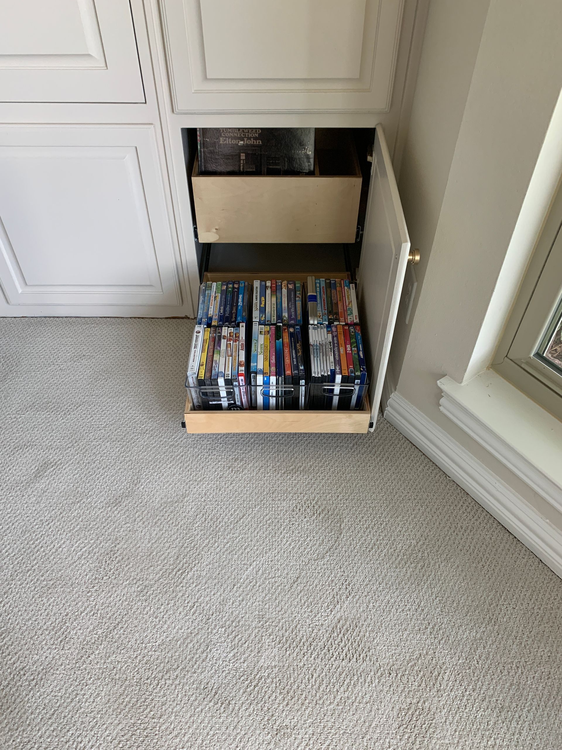 Pull-out cabinet with DVDs on a carpeted floor. White cabinets and a beige drawer.