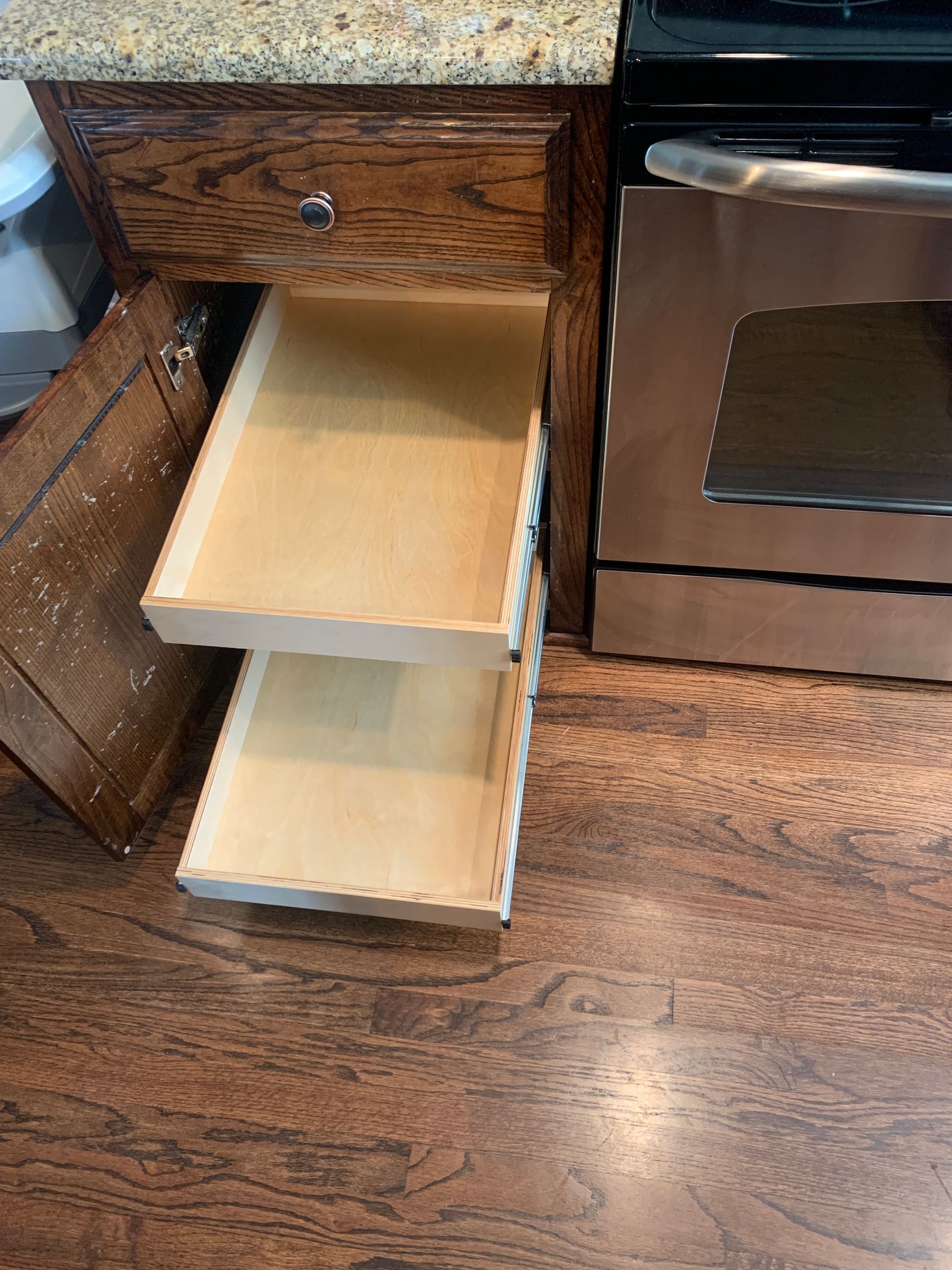 Two pull-out drawers in a kitchen cabinet, next to a stainless steel oven. Wooden floor.