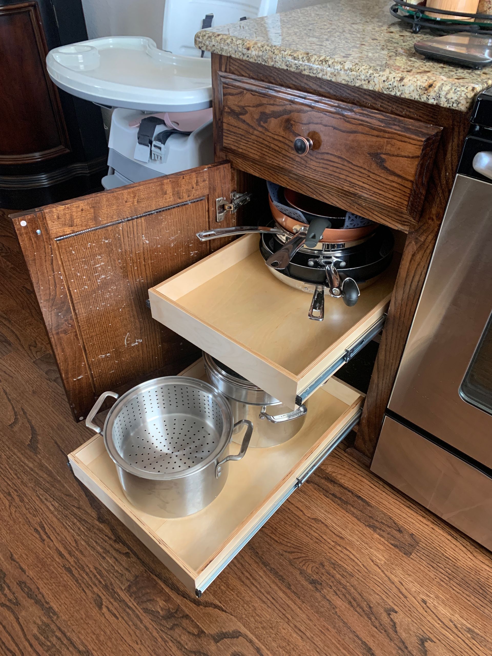 Kitchen cabinet with pull-out shelves holding cookware. Brown wood cabinet, stainless steel pots, and granite countertop.