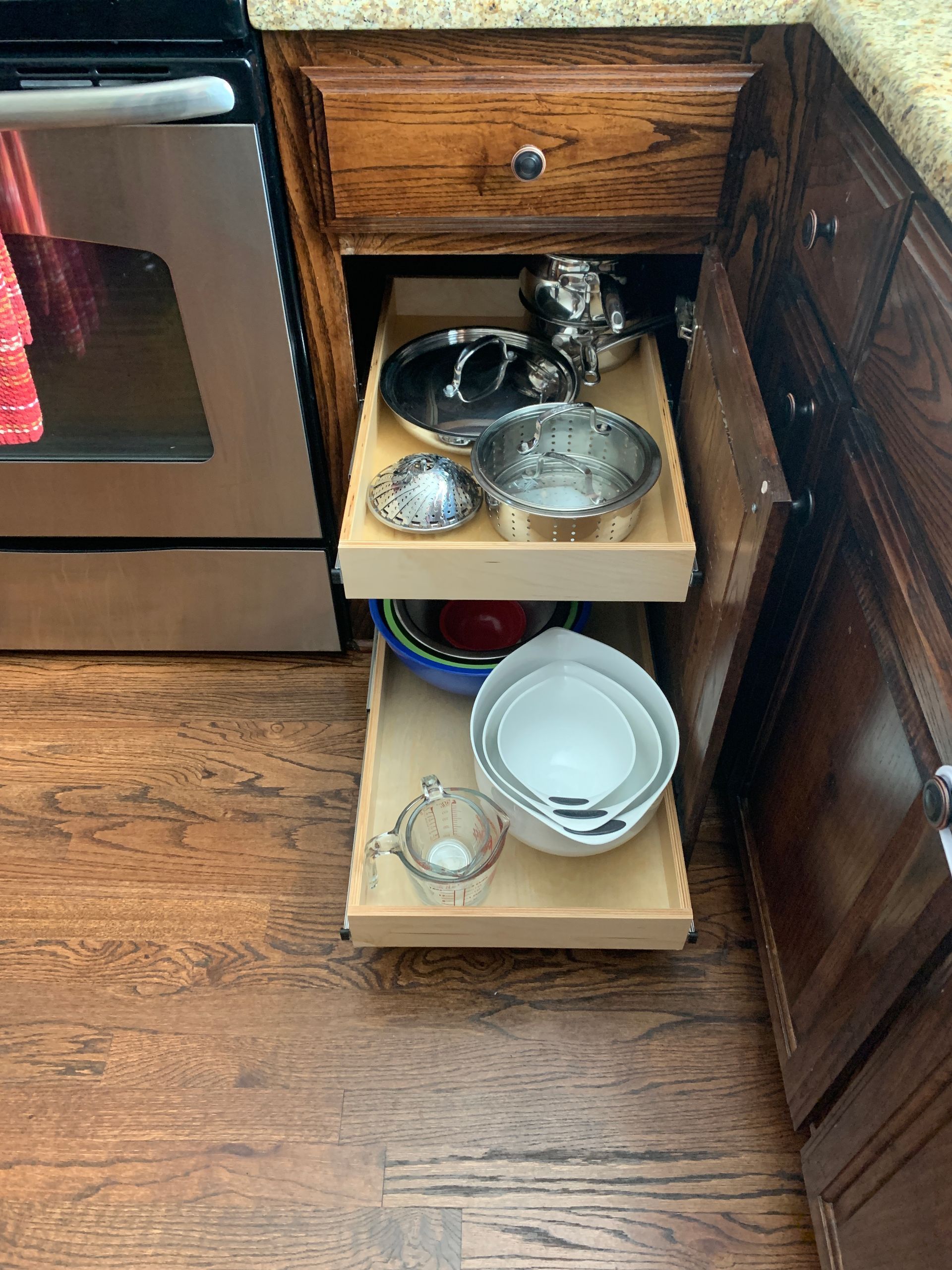 Pull-out cabinet drawers holding kitchen bowls, colander, and cookware. Oak cabinets, stainless steel stove, and wood floor.