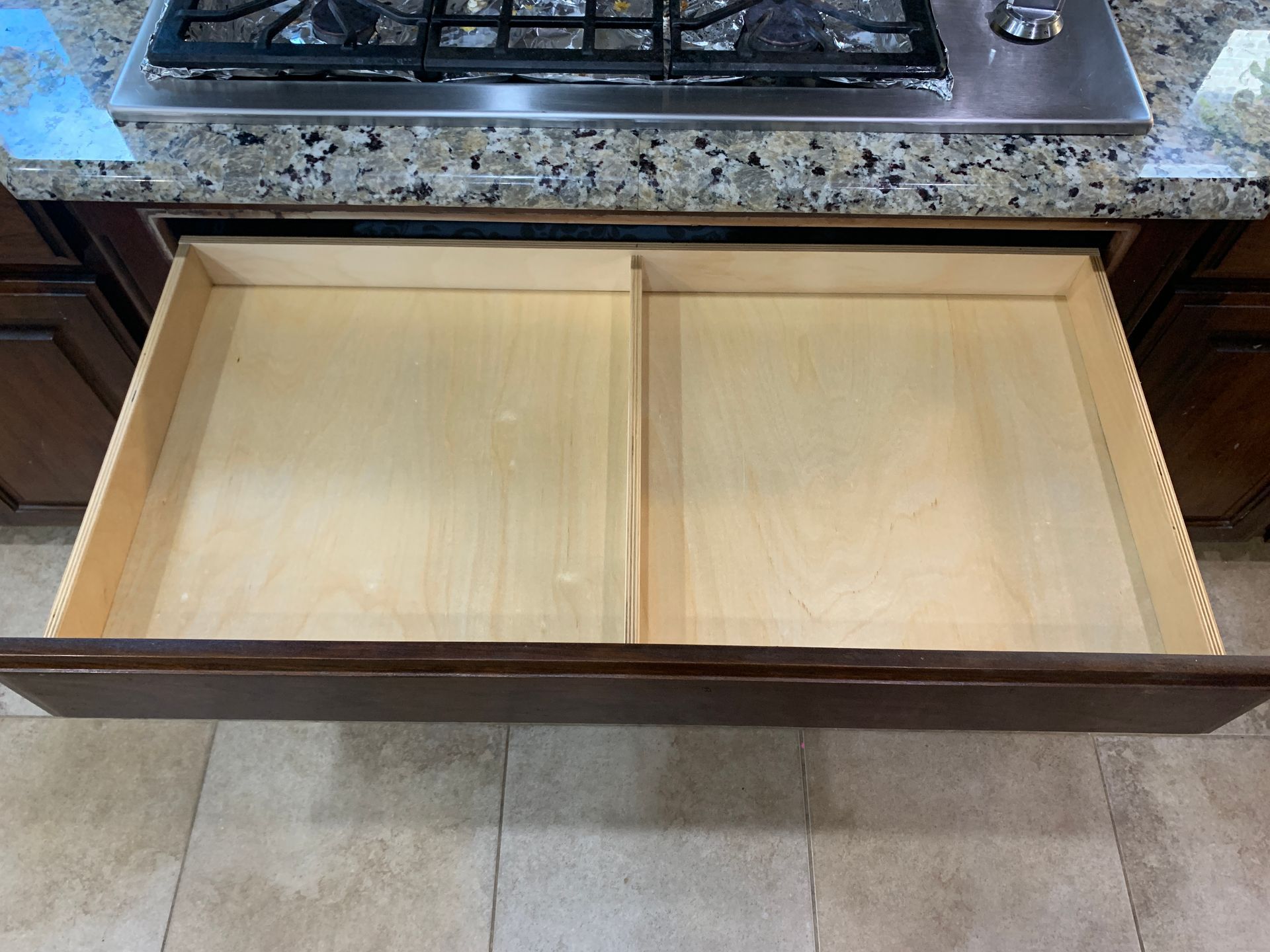 Open kitchen drawer with a central divider. Wooden interior, dark brown frame, below stovetop.