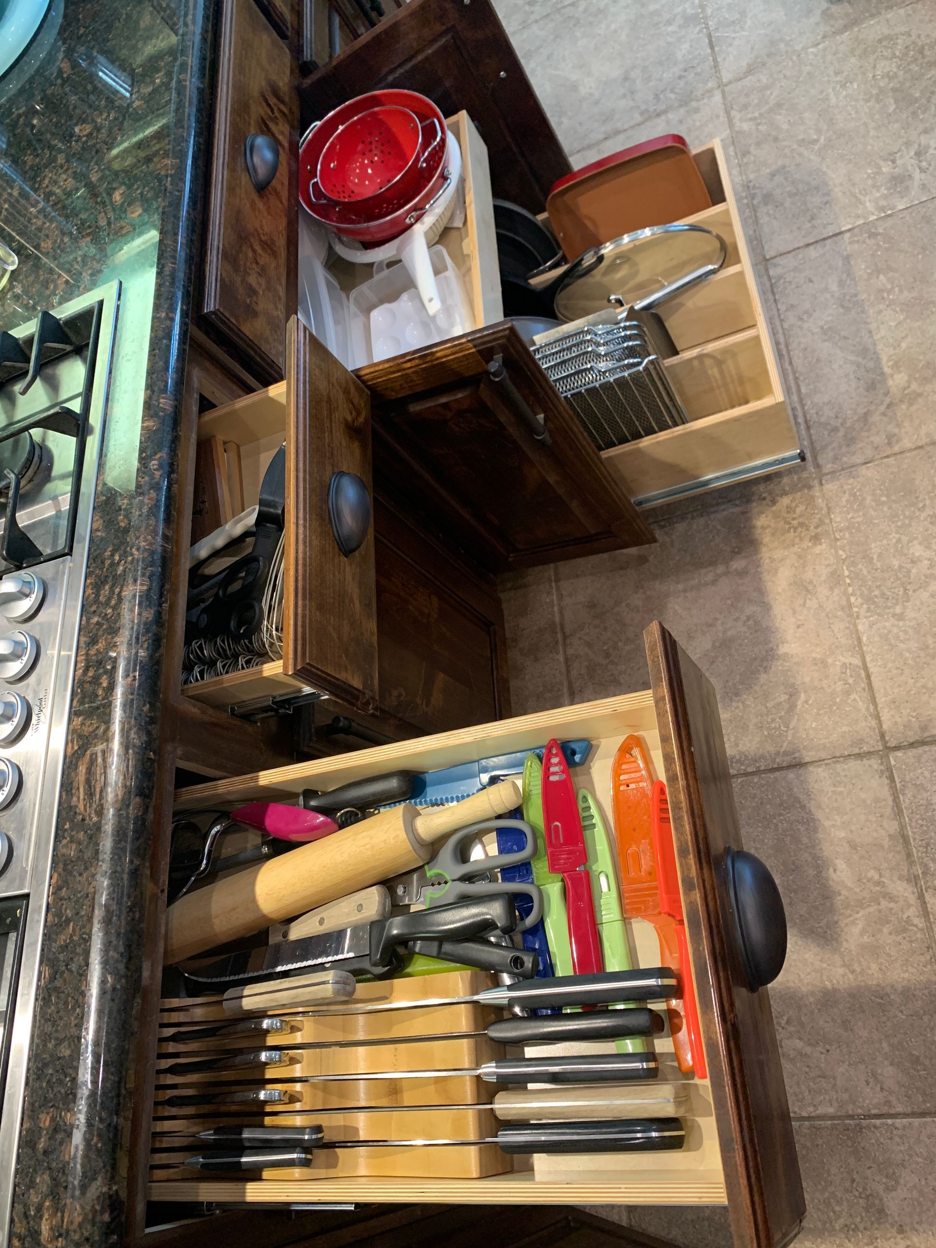 Open kitchen drawers holding cookware, utensils, and knives. Brown wood cabinets next to a stovetop.
