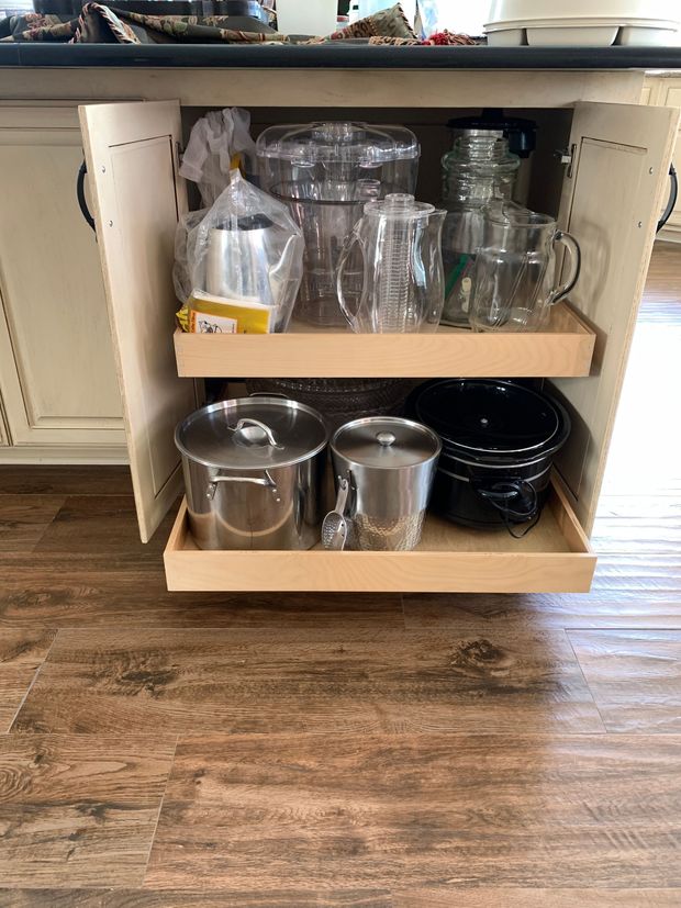 Kitchen cabinet with pull-out shelves holding glassware, pitchers, and a slow cooker. The cabinet is on a wood floor.
