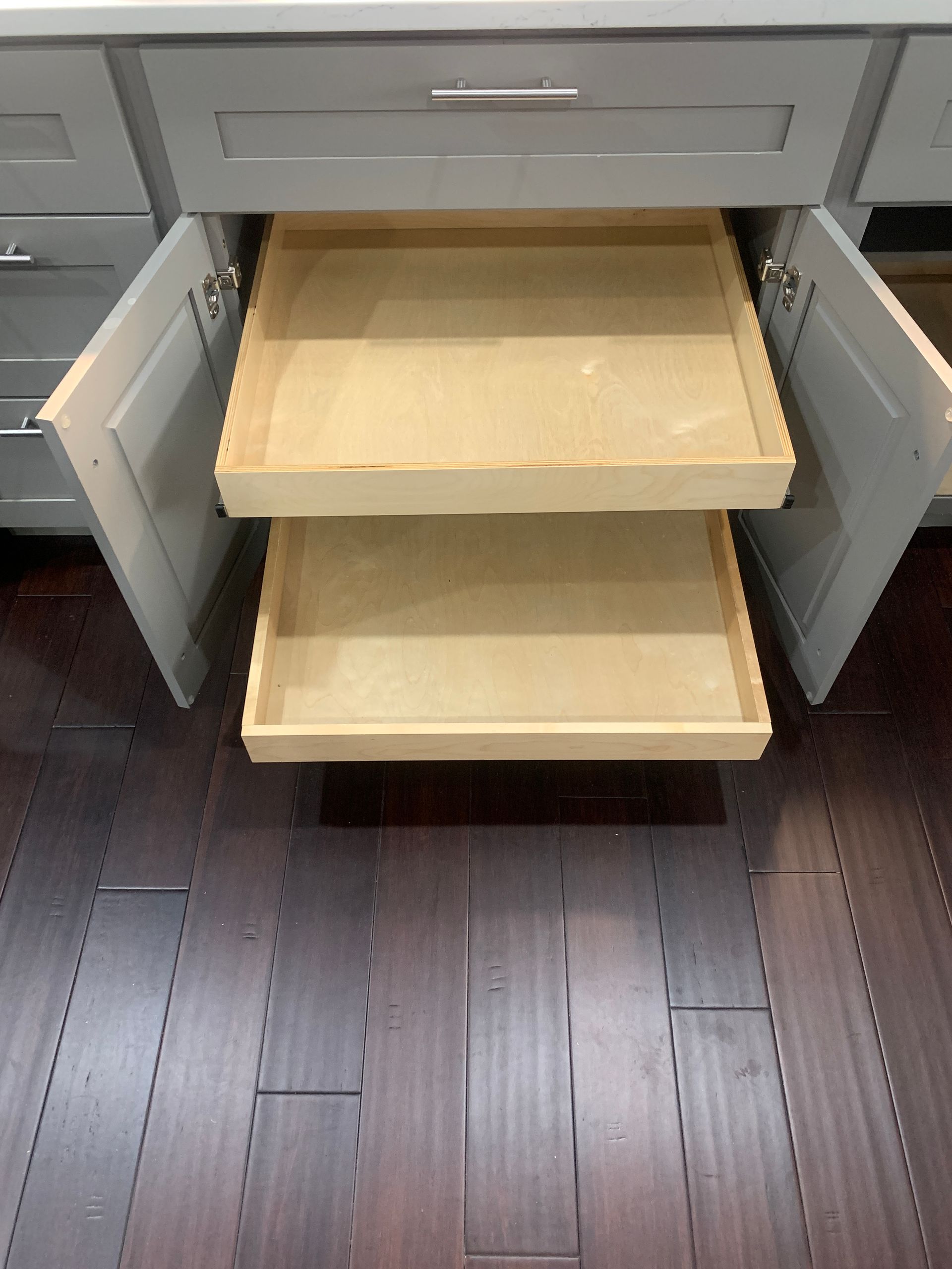 Two open pull-out shelves inside a gray kitchen cabinet. Dark wood floor.