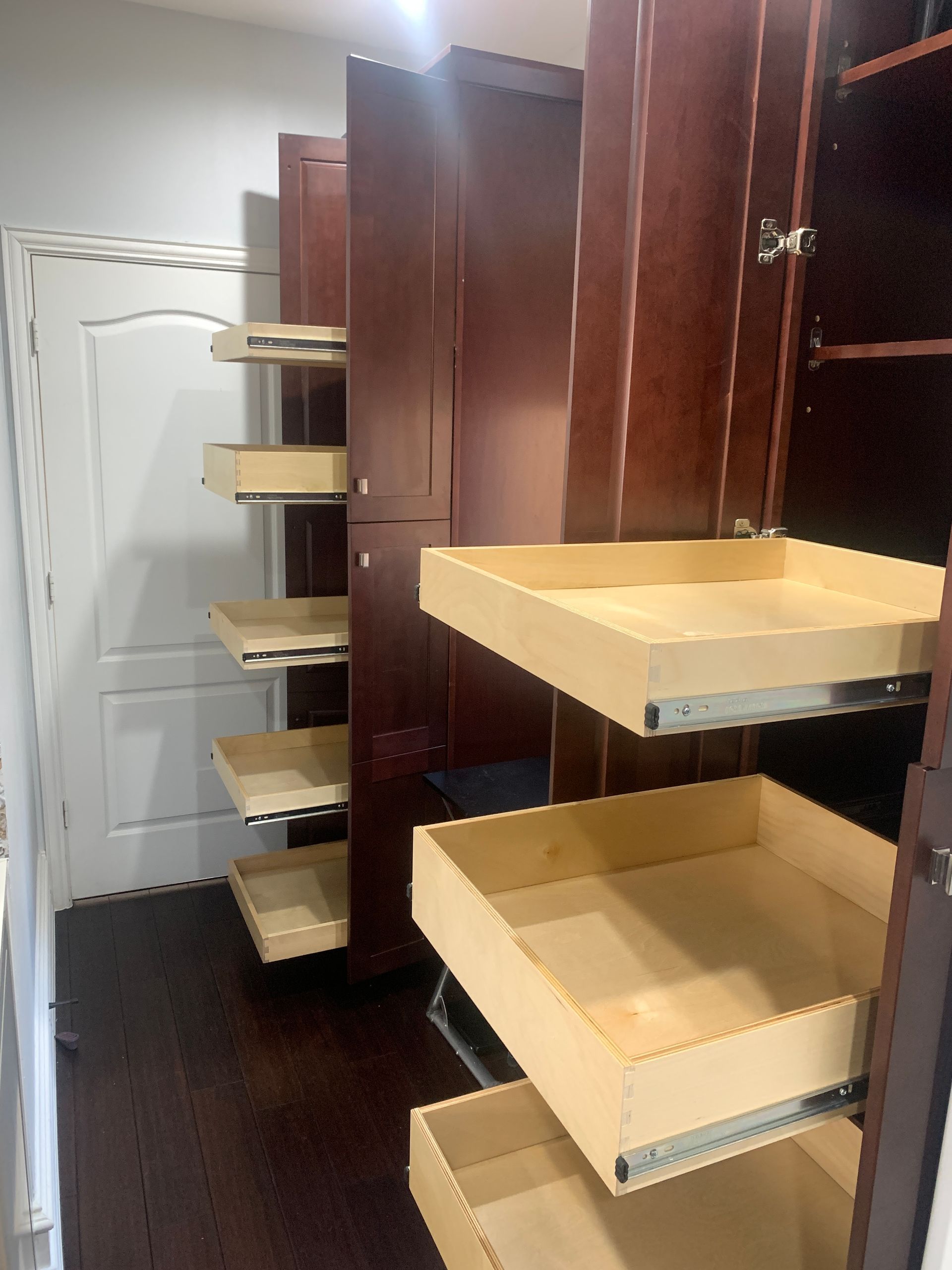 Wooden pull-out shelves inside tall, dark-brown cabinets in a room with hardwood floors and a white door.