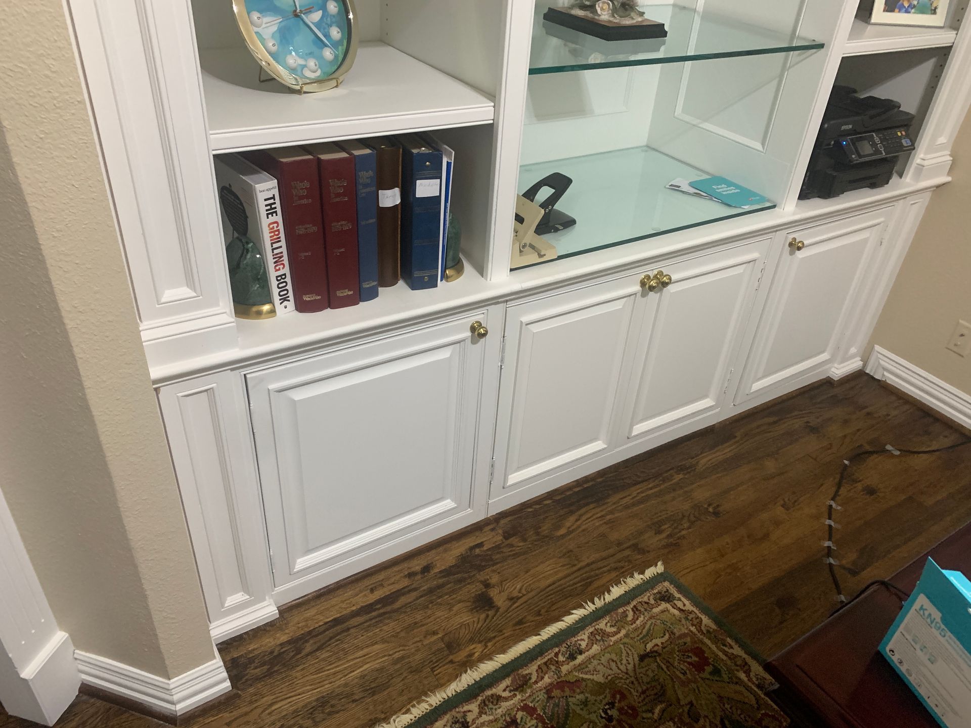 White built-in cabinet with glass shelves, books, and closed doors against a wood floor.