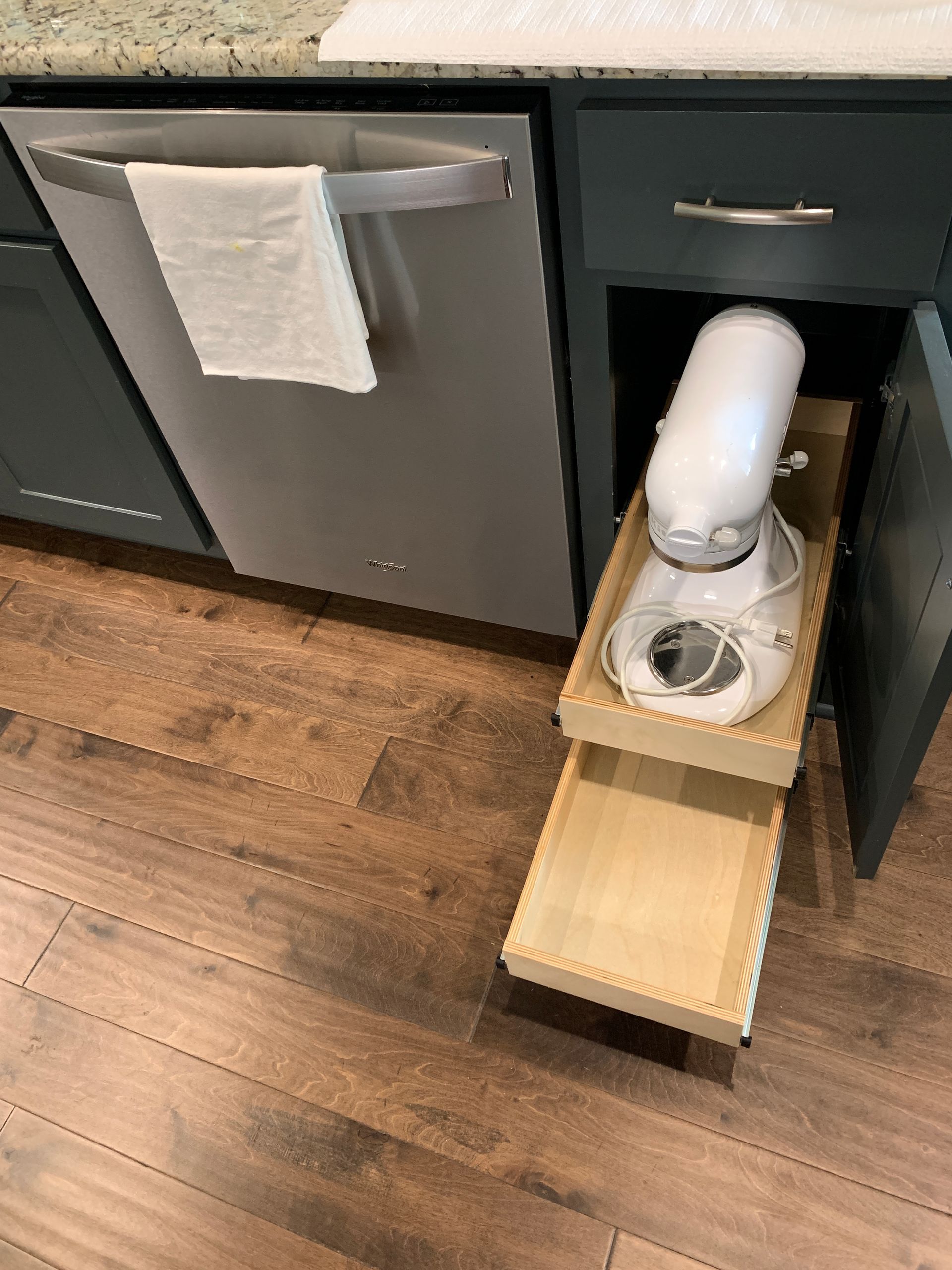 Kitchen cabinet with pull-out tray holding a white stand mixer. Next to a dishwasher, on a wood floor.