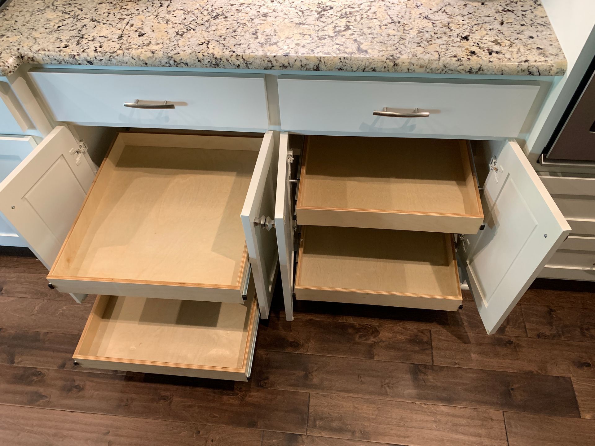 Open kitchen cabinets with pull-out drawers, white doors, granite countertop, and wood floor.