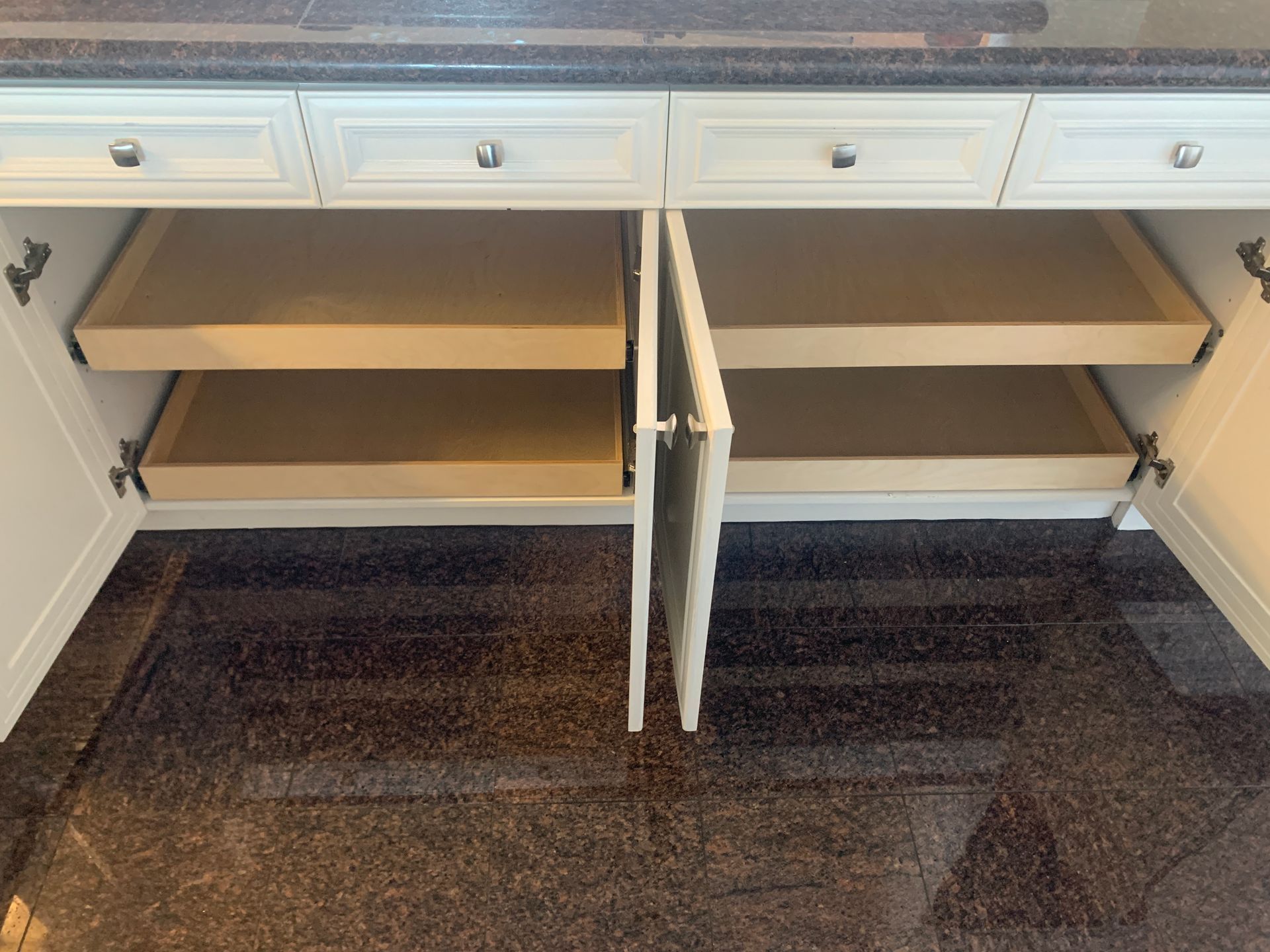 Open kitchen cabinet with pull-out shelves, cream-colored doors, dark countertop and flooring.