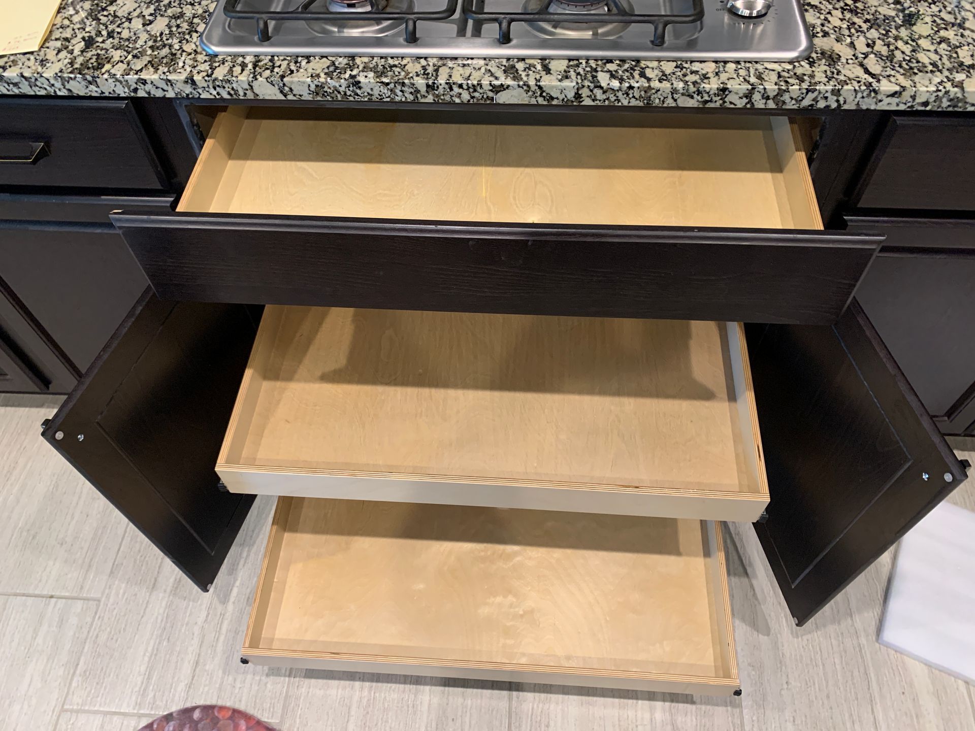 Open kitchen cabinet with three pull-out drawers, granite countertop, and dark brown cabinets.