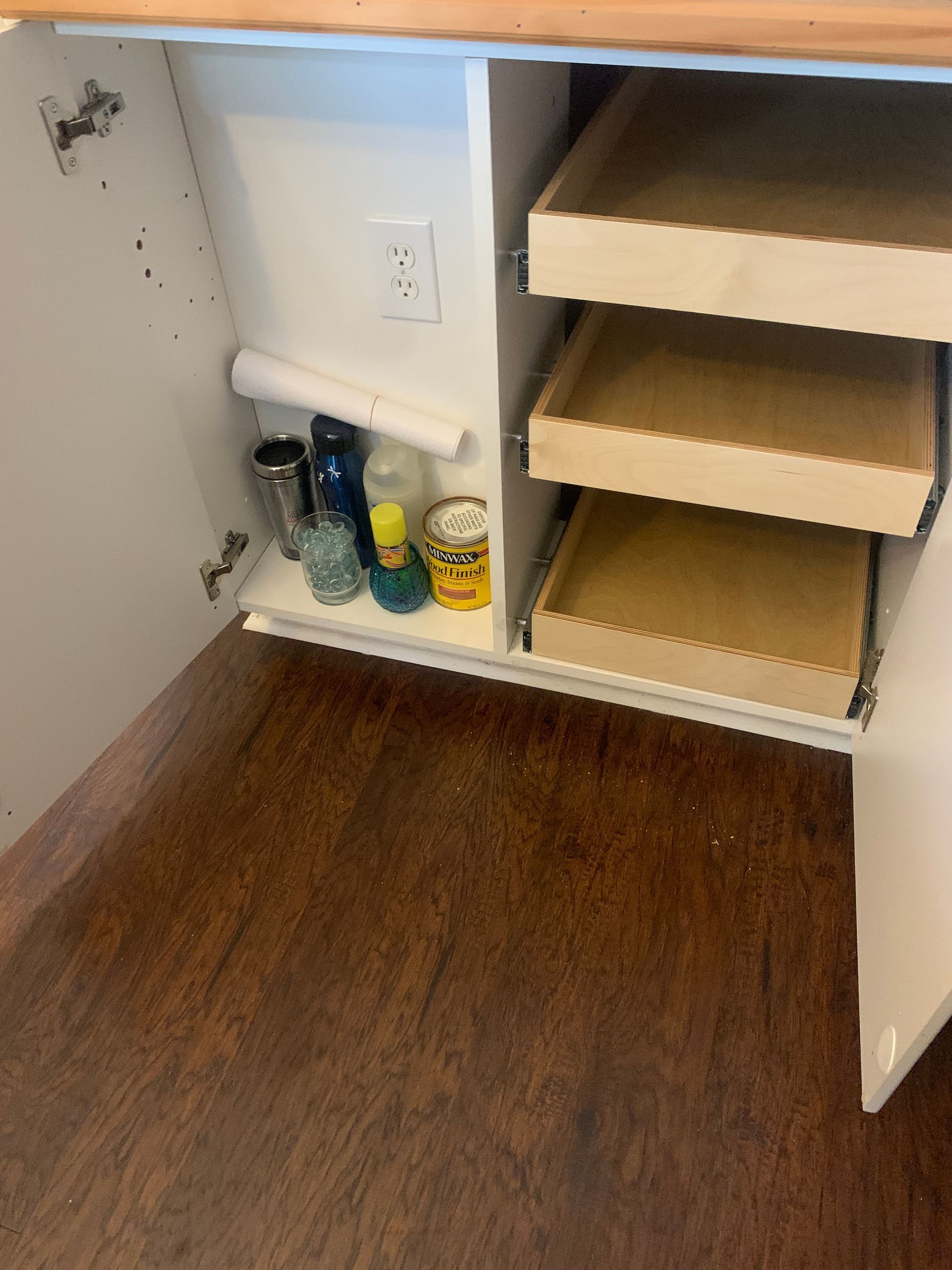 Cabinet interior with pull-out shelves and stored items, including bottles and a paper towel roll. Brown floor.