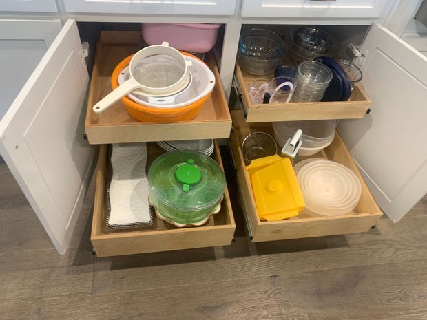 Open kitchen cabinet with pull-out drawers holding various kitchen items, including bowls, strainers, and a salad spinner.