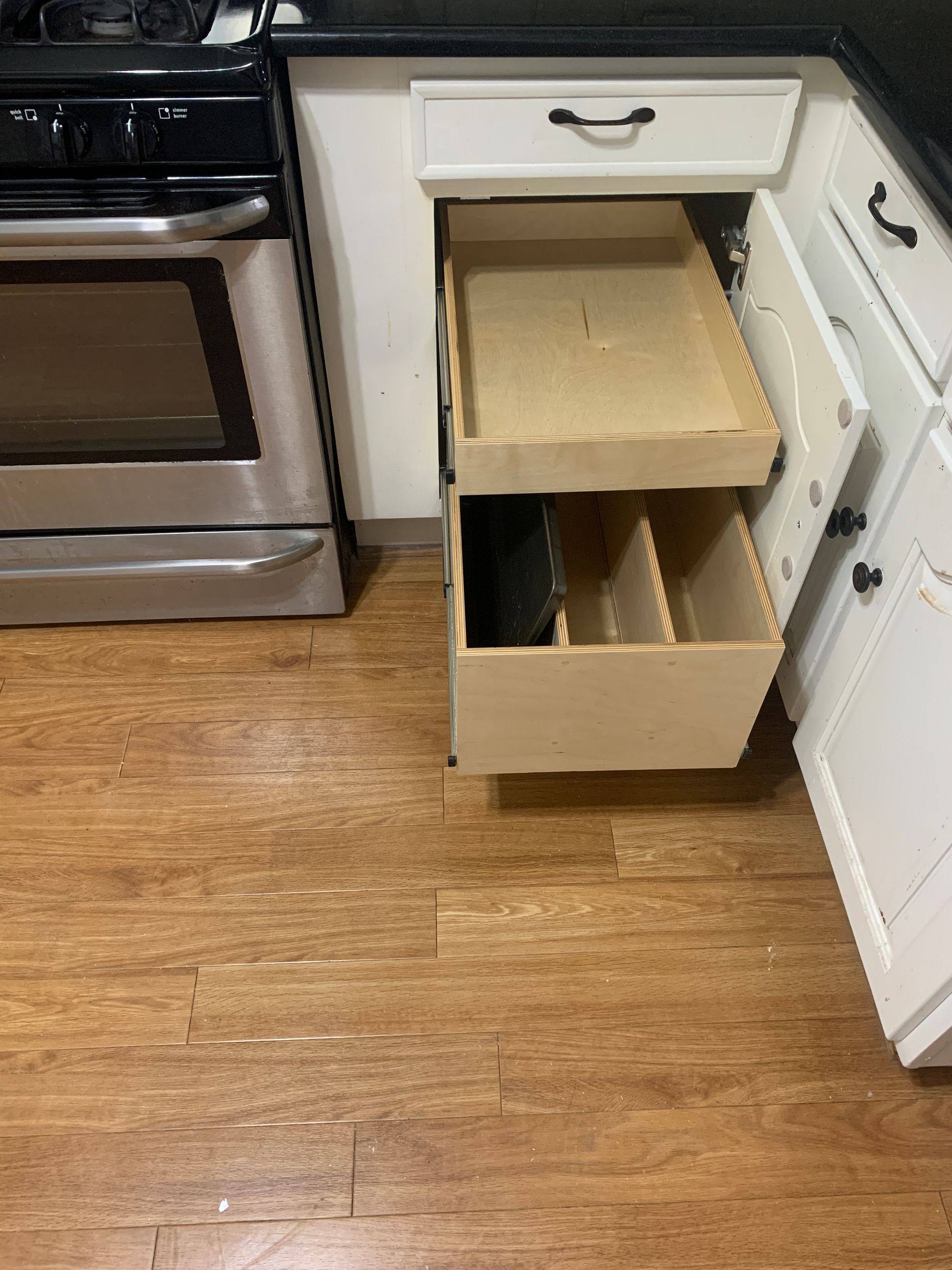Kitchen cabinet with pull-out drawers, next to a stove. Wooden floor. White cabinets.