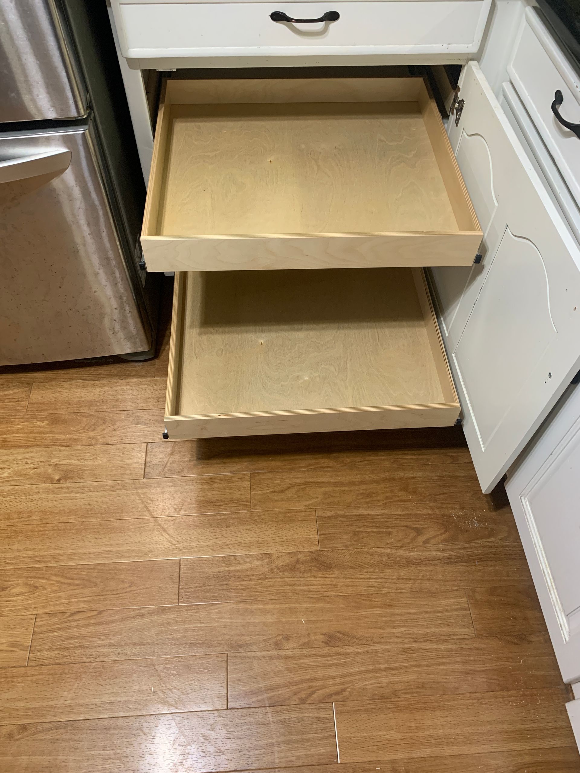 Two pull-out shelves in a white kitchen cabinet are open. The shelves are empty, made of light wood.