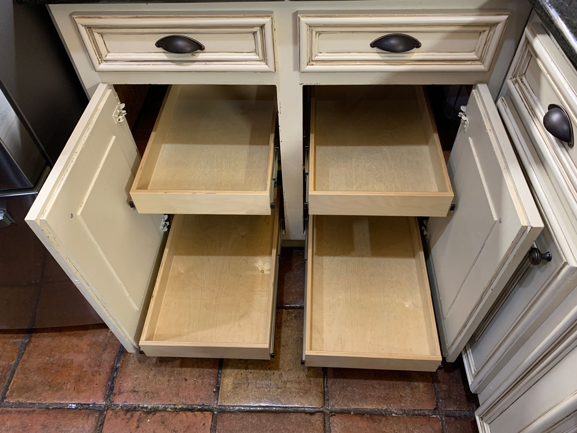 Open kitchen cabinet with pull-out drawers, cream-colored doors and trim, and black hardware.