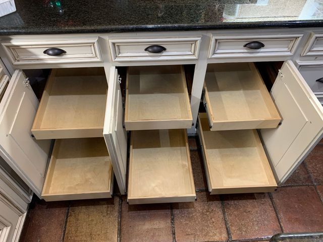Cream-colored kitchen cabinets with open drawers revealing pull-out shelves. Dark countertop and tile floor.