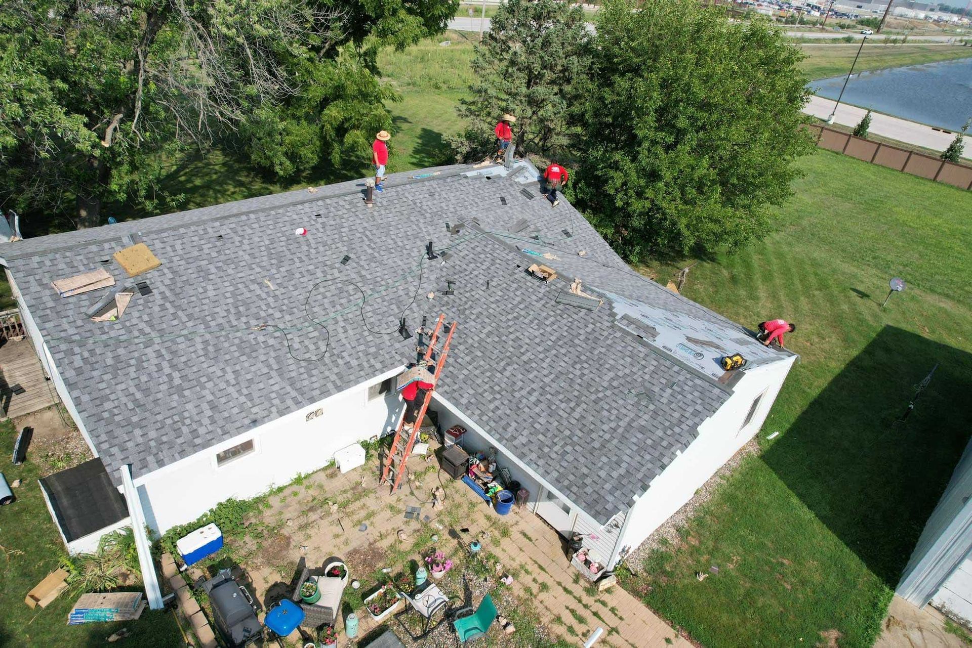 Workers on a gray shingled roof, repairing it beside a white house and green yard.