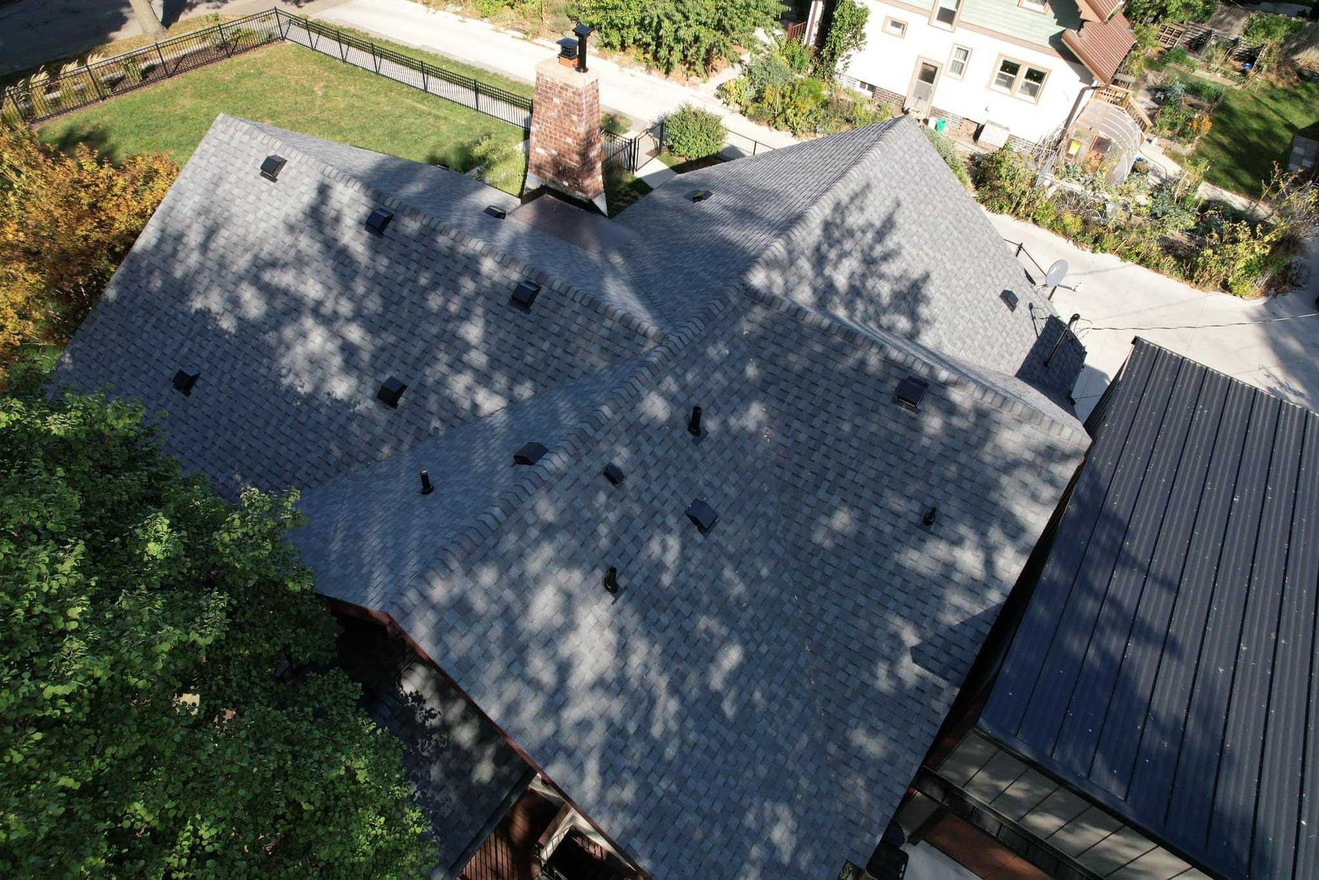 Aerial view of a house roof with a central chimney, surrounded by trees and neighboring homes.
