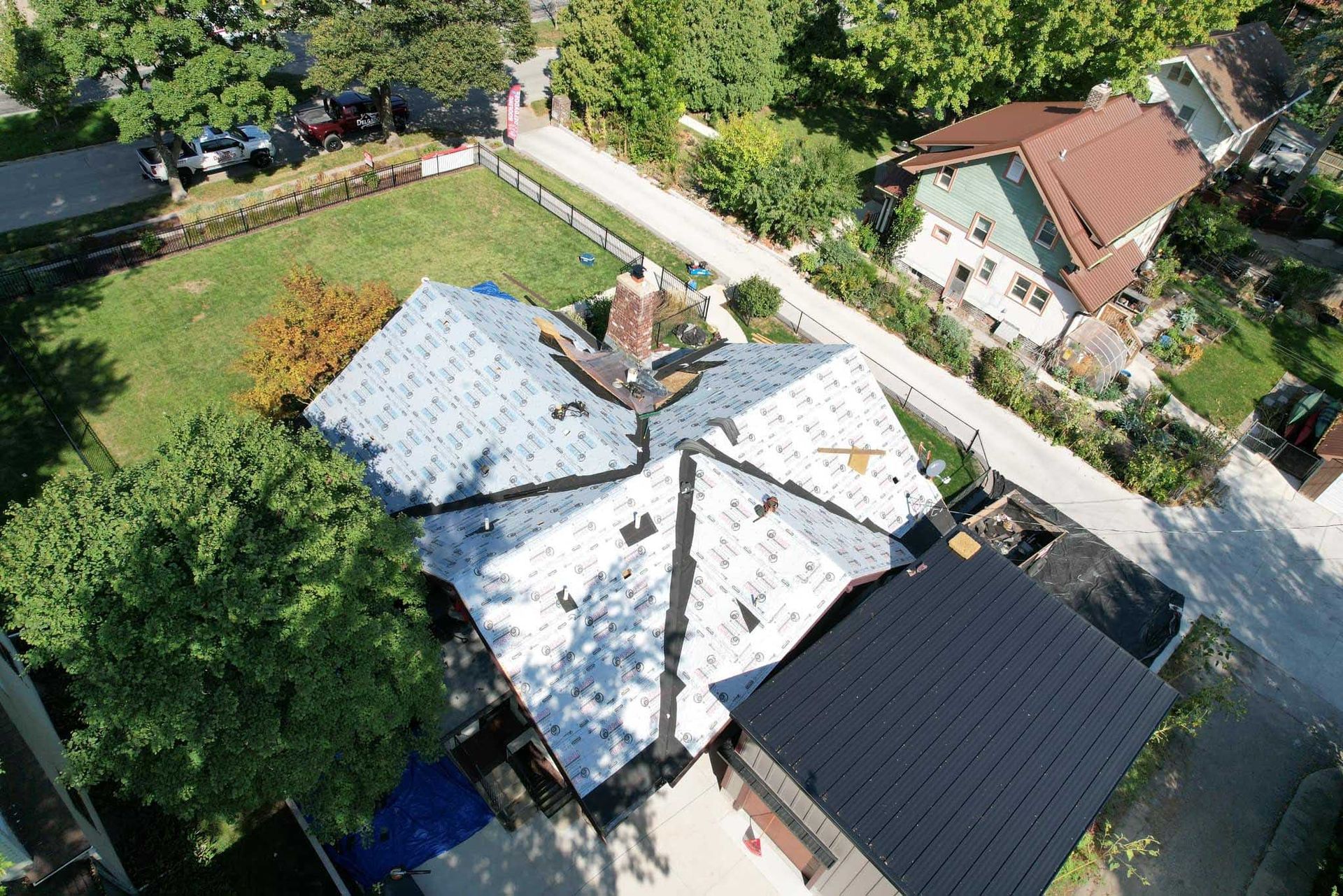 Aerial view of a house with a white roof beside a road, trees, and neighboring buildings.